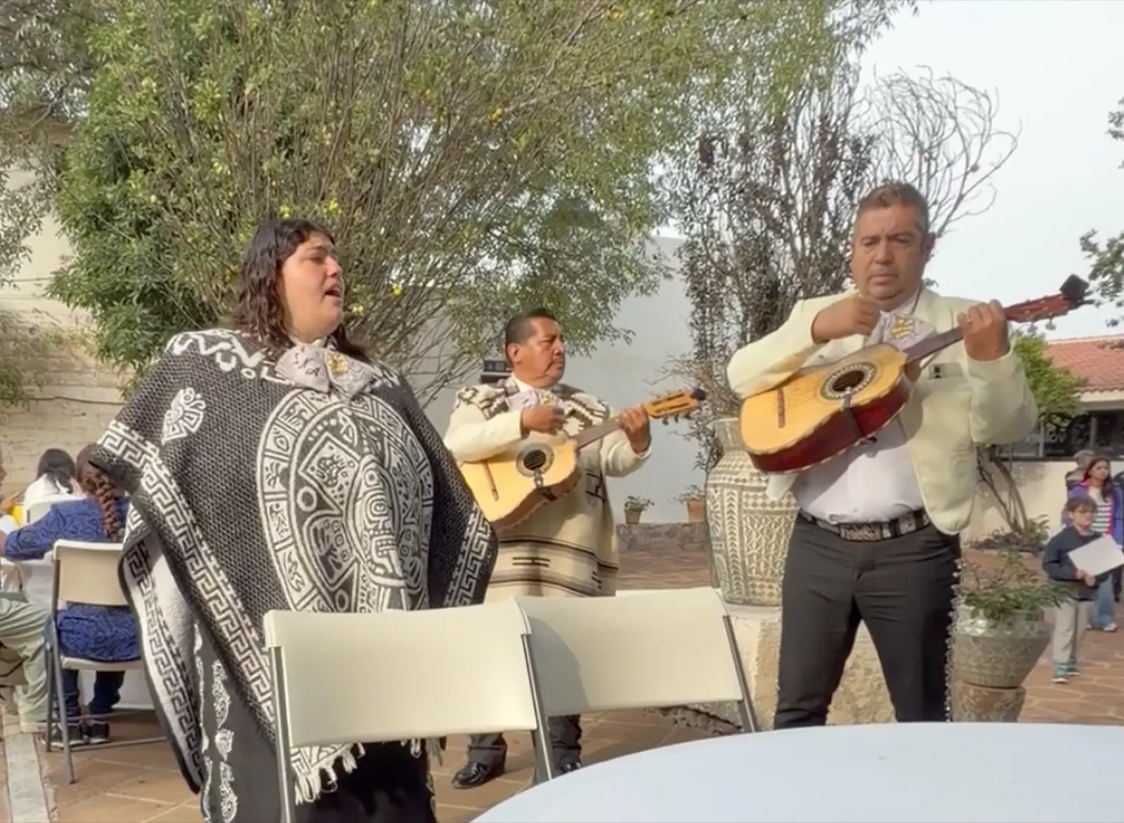 Three mariachi musicians in traditional dress performing outdoors, one singing and two playing guitars
