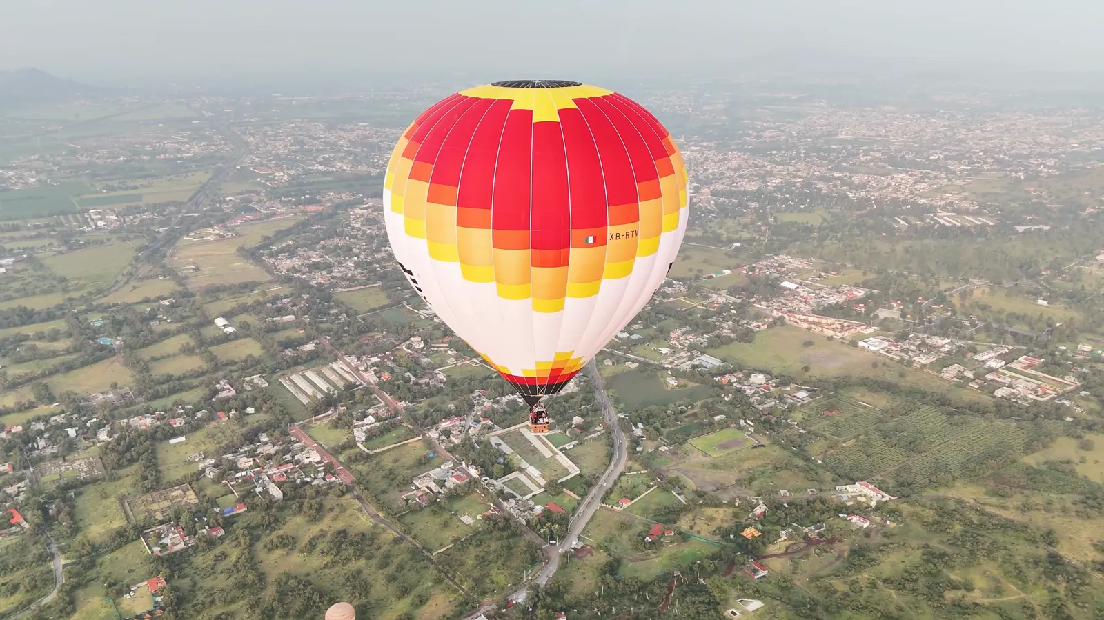 Drone view of a red and yellow hot air balloon drifting over farmland and rooftops
