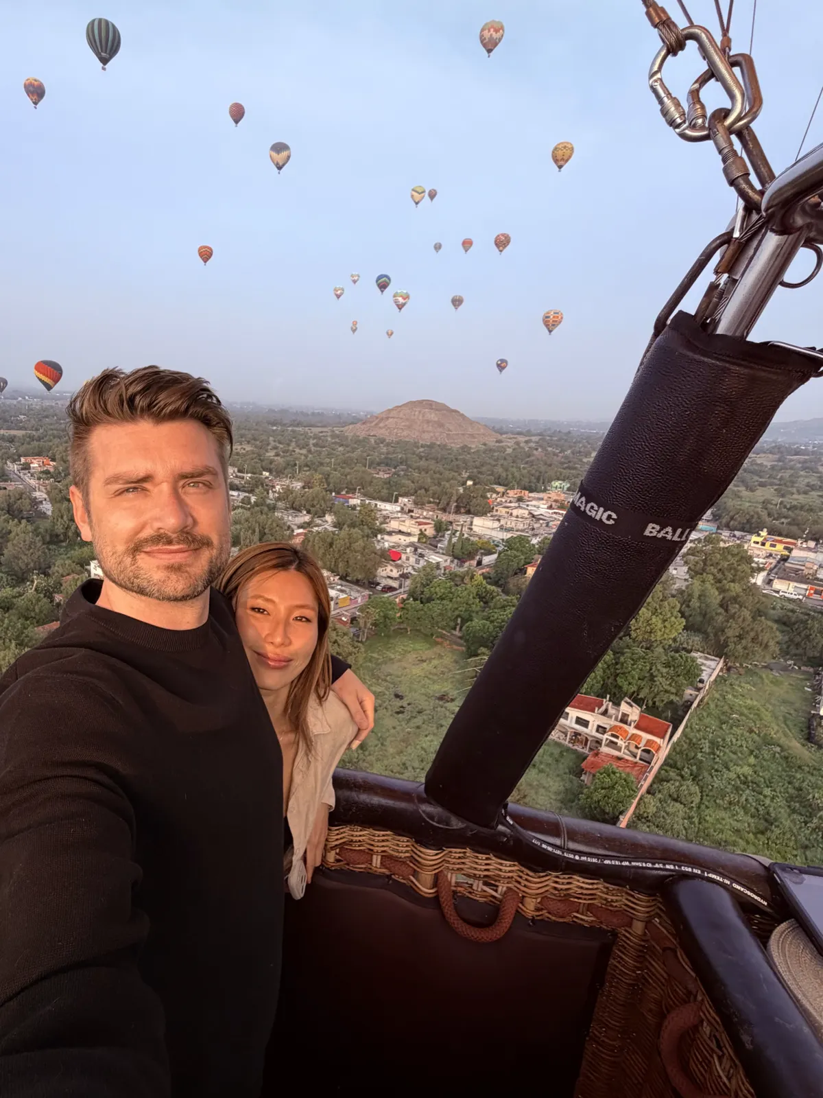 Couple in a hot air balloon basket at sunrise with dozens of balloons in the sky and the pyramid below