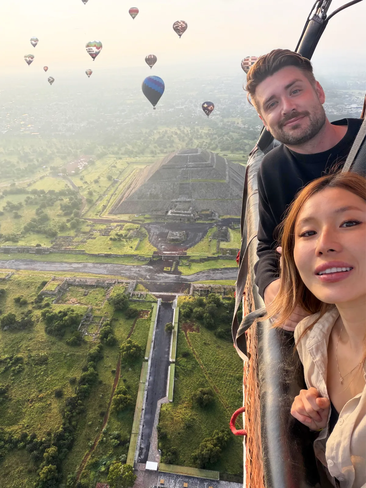 Couple leaning out of a hot air balloon basket at sunrise with the Teotihuacan pyramid complex and dozens of balloons below