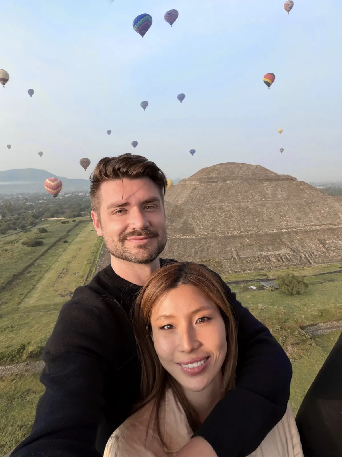 Couple selfie from a balloon basket with the Pyramid of the Sun behind them and dozens of balloons in the sky