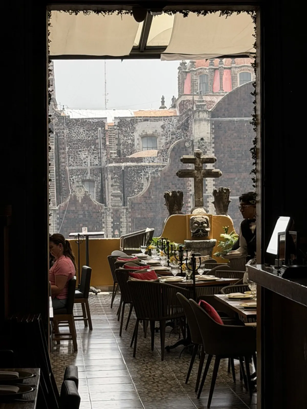 Looking out through a dark doorway onto a sunlit rooftop restaurant terrace with stone church towers beyond