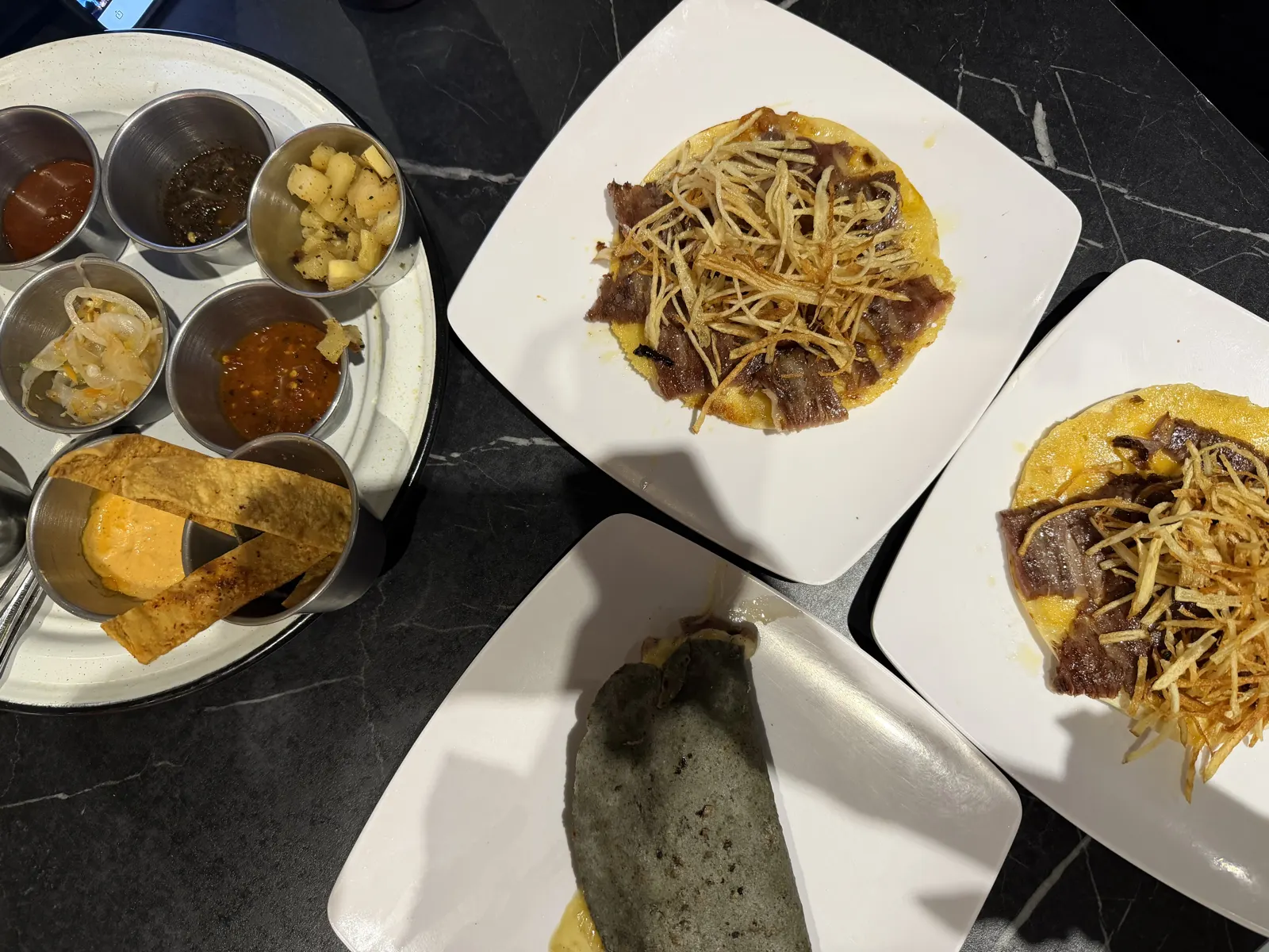 Two plates of steak tacos with shoestring potatoes beside a tray of six salsas and condiments